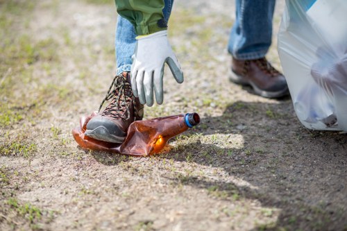 A close-up of a person wearing blue jeans, a green jacket, and white gloves, picking up a crushed brown glass bottle from a patch of ground in an outdoor setting. The bottle has a blue neck and cap, and is partially resting on a gravel surface mixed with dirt. In the background, another person wearing dark brown shoes and blue jeans is partially visible holding a large transparent plastic bag filled with other rubbish, typical of waste collection or rubbish removal activities in an outdoor space. The scene suggests a cleanup operation, consistent with services provided by Garden Clearance Hayes, focused on garden or outdoor rubbish removal in the Hayes area of West London, with a clear emphasis on waste materials such as glass bottles and general refuse commonly encountered during clearance work.